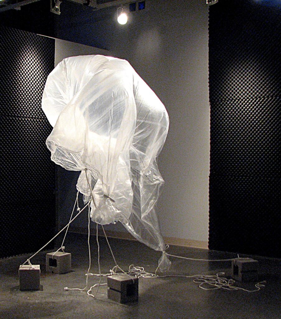 Four cinder blocks hold down a transparent, baloon-like circle of plastic. Light shines down on it in a room that has gray and black walls.